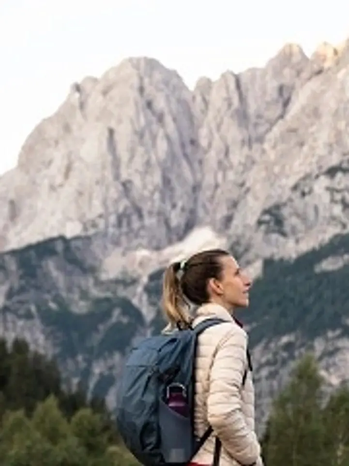 rs=w_721,h_961,cg_true A woman with a backpack is standing in front of some mountains.